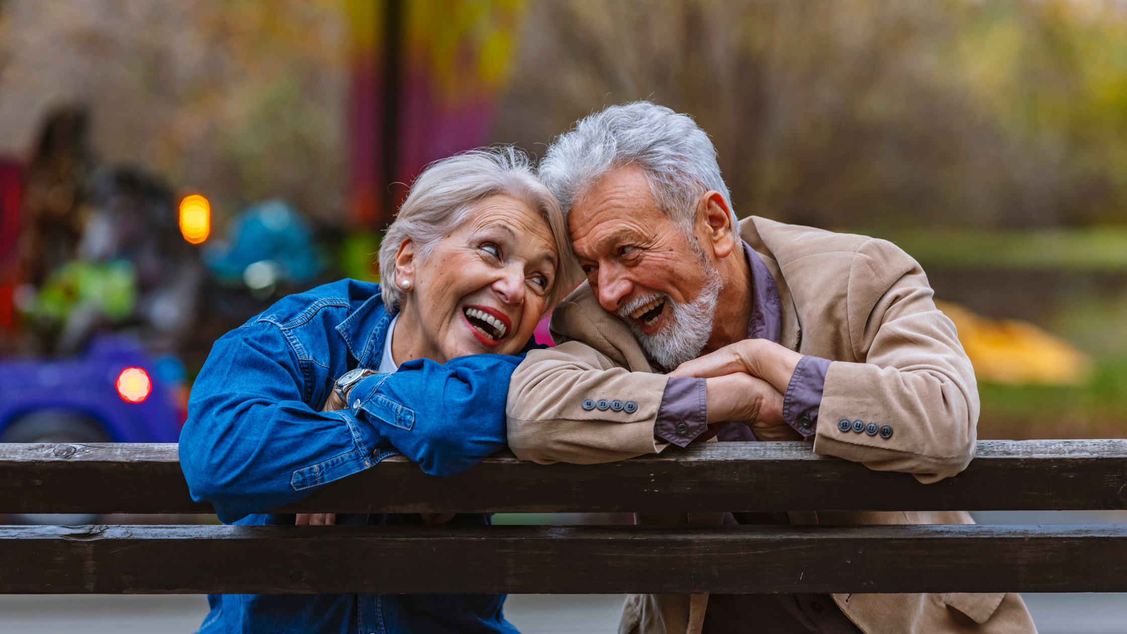 Retired couple looking at each other laughing in the park on a bench.